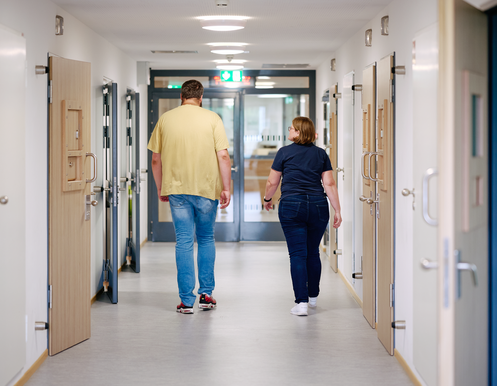 Eine Mitarbeiterin und ein Patient laufen einen Stationsflur entlang.